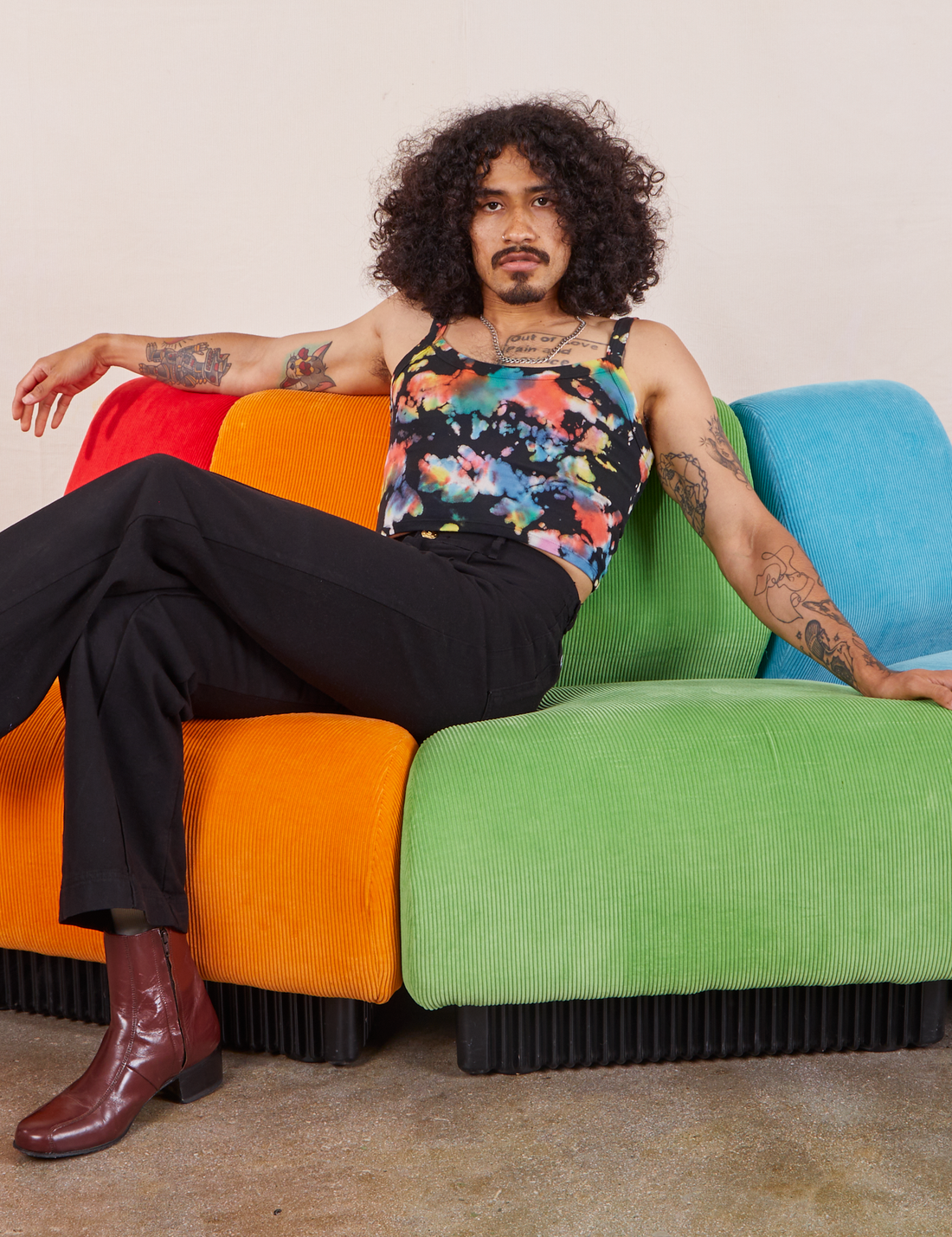 Jesse is sitting on an upholstered chair wearing Cropped Cami in Rainbow Magic Waters and black Western Pants