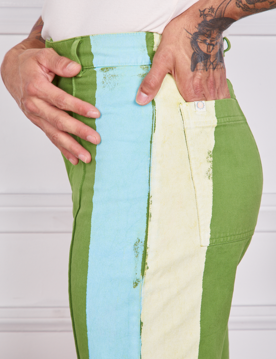 Side close up of Hand-Painted Stripe Western Pants in Bright Olive. Baby blue and off-white hand painted stripe along the side. Jesse has their hand in the back pocket.