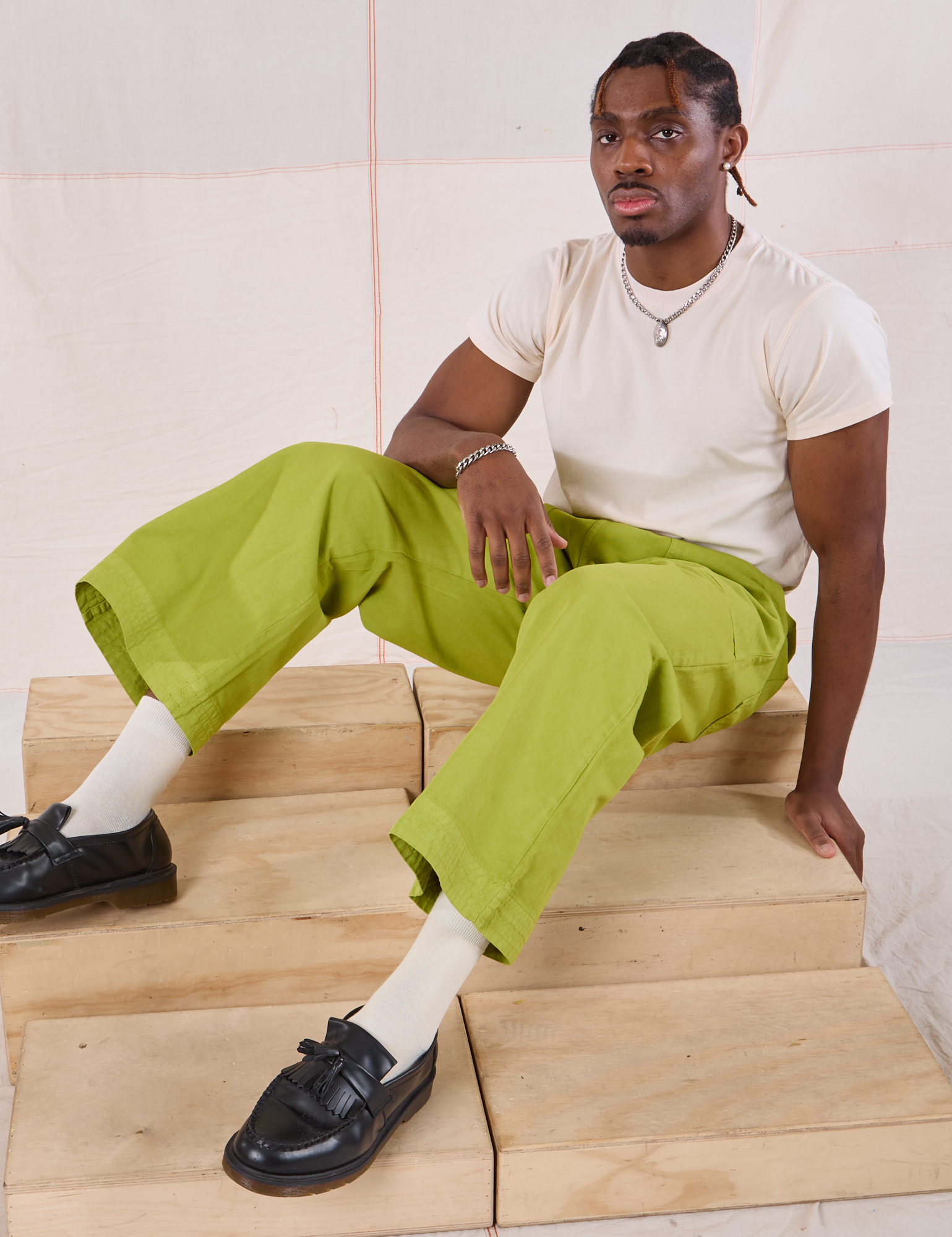 Isaac is wearing Action Pants in Gross Green and sitting on a wooden crate