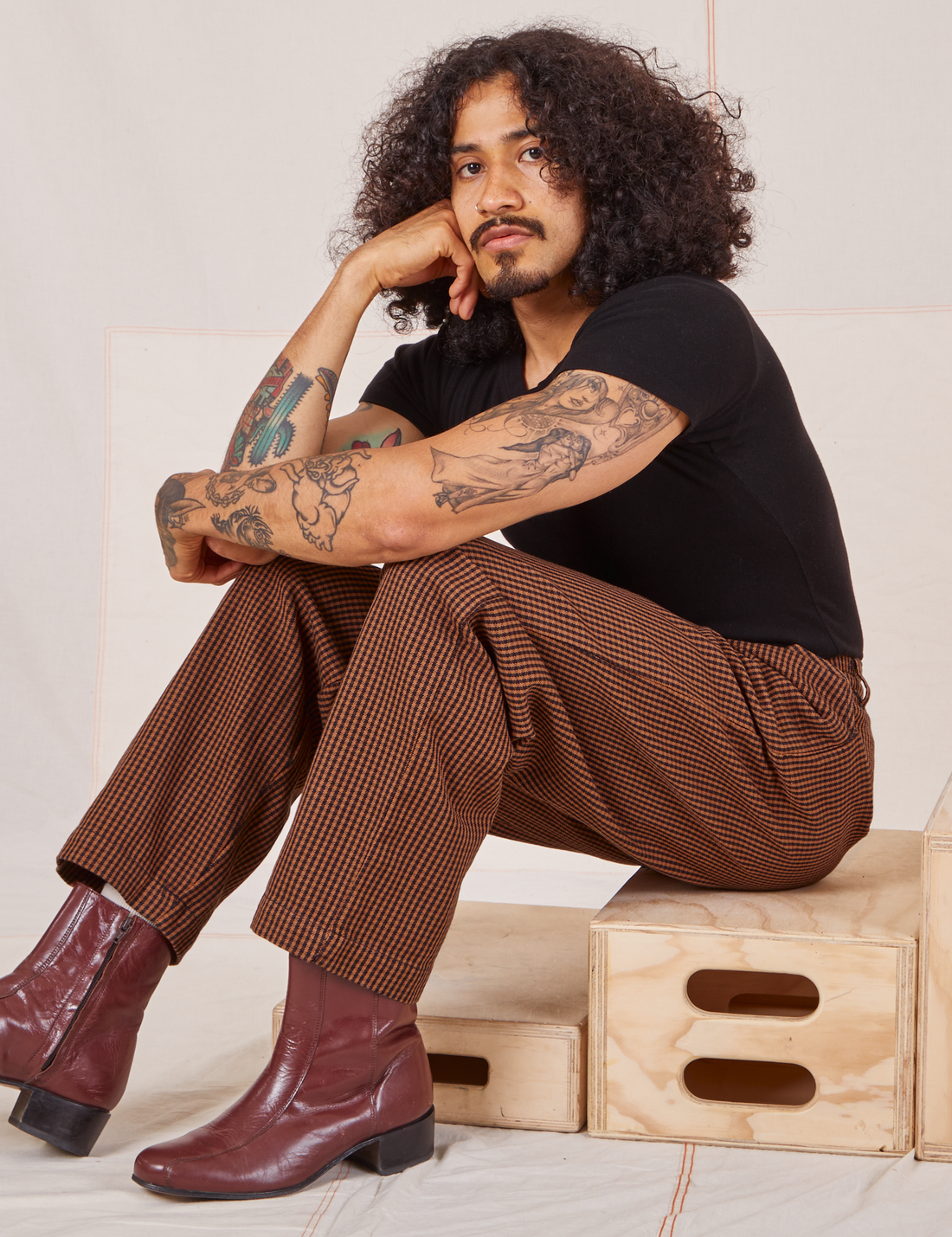 Jesse is sitting on a wooden crate. They are wearing Checker Trousers in Brown and black Baby Tee