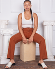 Gabi is sitting on a wooden crate wearing Hand-Painted Stripe Western Pants in Burnt Terracotta and a vintage off-white Tank Top