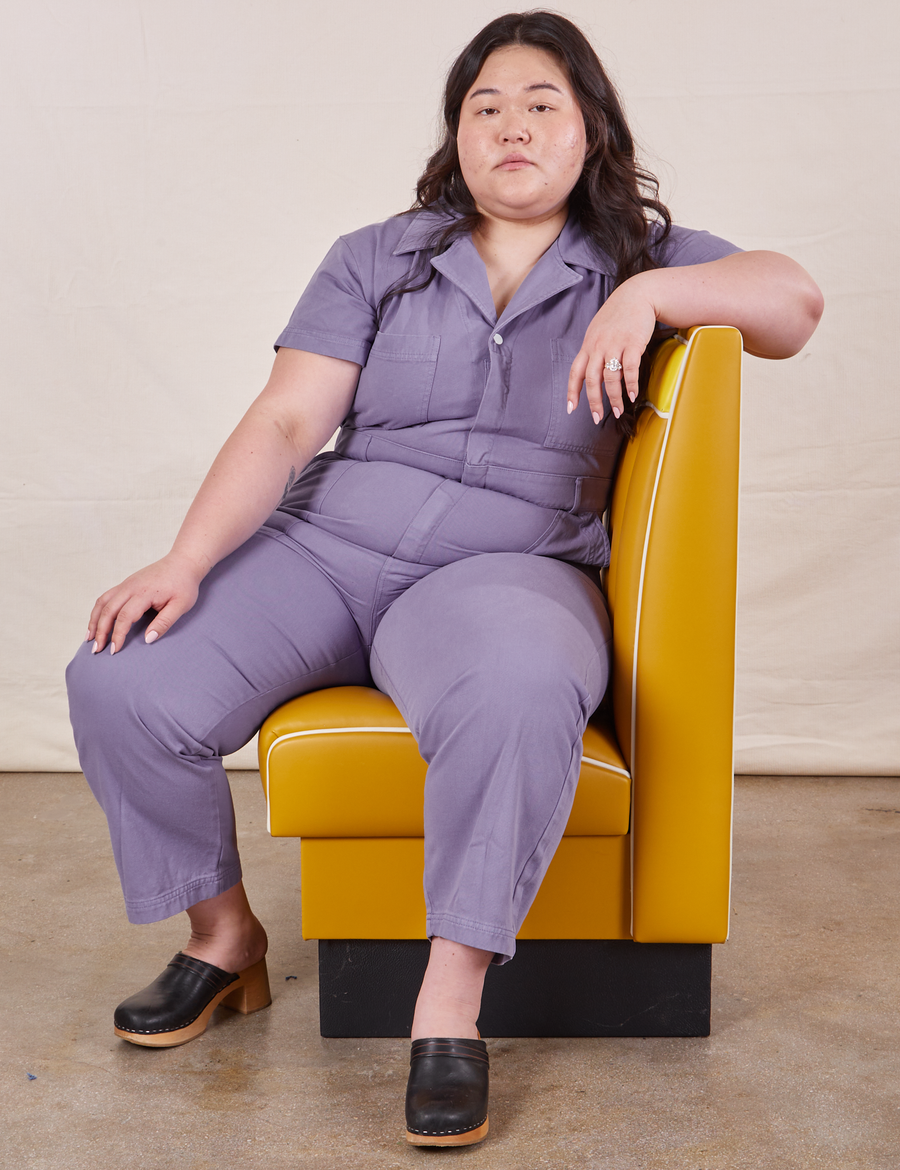 Ashley is sitting in a mustard yellow vinyl booth seat wearing Petite Short Sleeve Jumpsuit in Faded Grape