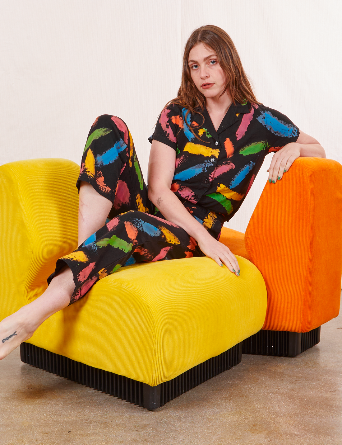 Allison is sitting on a yellow upholstered chair wearing Pantry Button Up in Paint Stroke and matching Work Pants
