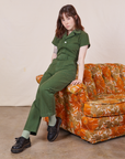 Hana is sitting on the armchair of a sofa wearing Petite Short Sleeve Jumpsuit in Dark Emerald Green