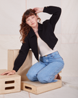 Alex is kneeling on a wooden crate wearing Ricky Jacket in Basic Black