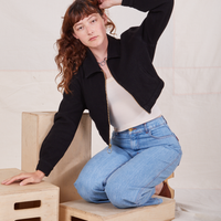 Alex is kneeling on a wooden crate wearing Ricky Jacket in Basic Black