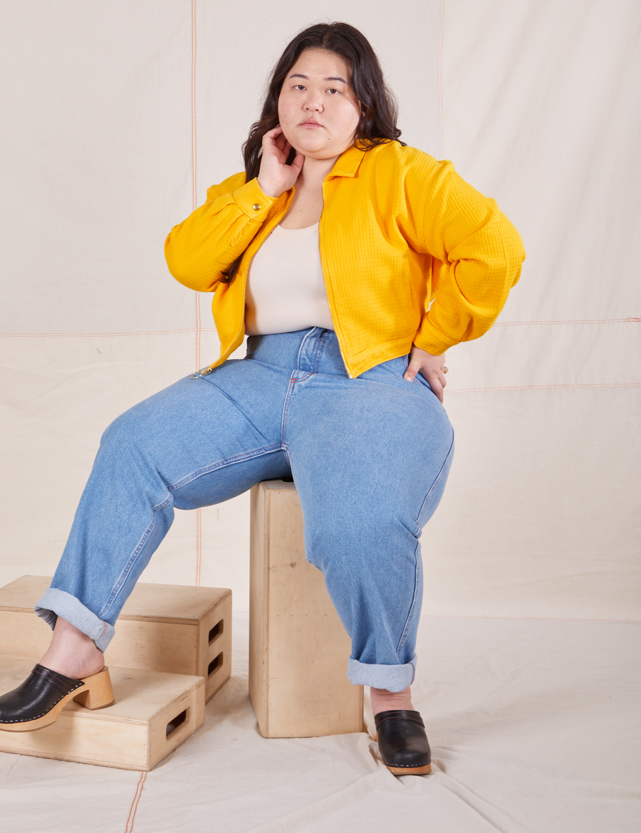 Ashley is sitting on a wooden crate wearing the Ricky Jacket in Sunshine Yellow, a vintage off-white Tank Top and light wash Frontier Jeans