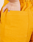 Back pocket close up of Short Sleeve Jumpsuit in Mustard Yellow. Alex has her hand in the pocket.