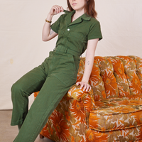 Hana is sitting on the arm of a sofa wearing Petite Short Sleeve Jumpsuit in Dark Emerald Green and holding the collar