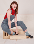 Alex is sitting on a wooden crate wearing Railroad Stripe Denim Original Overalls and paprika Sleeveless Turtleneck
