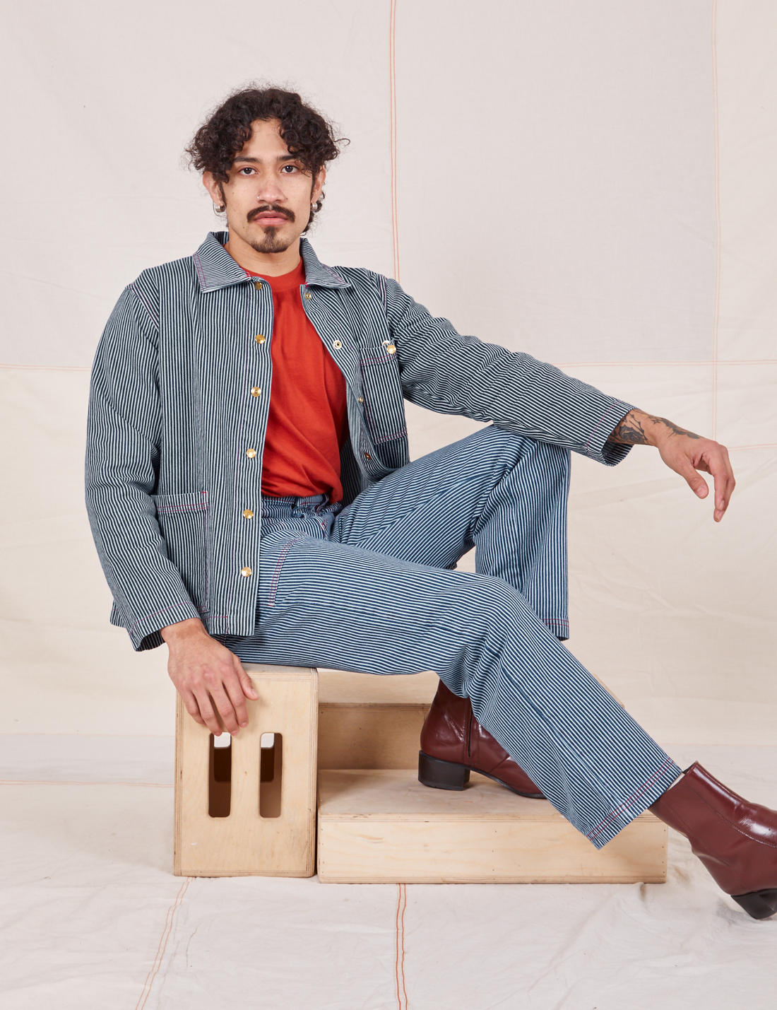 Jesse is sitting sideways on a wooden crate wearing Railroad Stripe Denim Work Jacket paired with matching Work Pants
