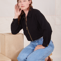 Alex is kneeling on a wooden crate wearing Ricky Jacket in Basic Black and light wash Sailor Jeans
