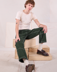 Quinn is sitting on a stack of wooden crates in his Carpenter Jeans in Swamp Green