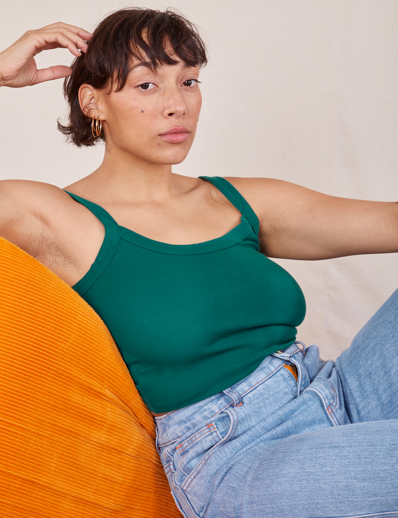 Tiara is sitting on a orange upholstered chair wearing Cropped Cami in Hunter Green and light wash Sailor Jeans