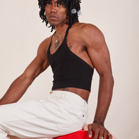 Jerrod is sitting on a red circular platform with his body facing the left side. They are wearing Halter Top in Basic Black and vintage off-white Western Pants
