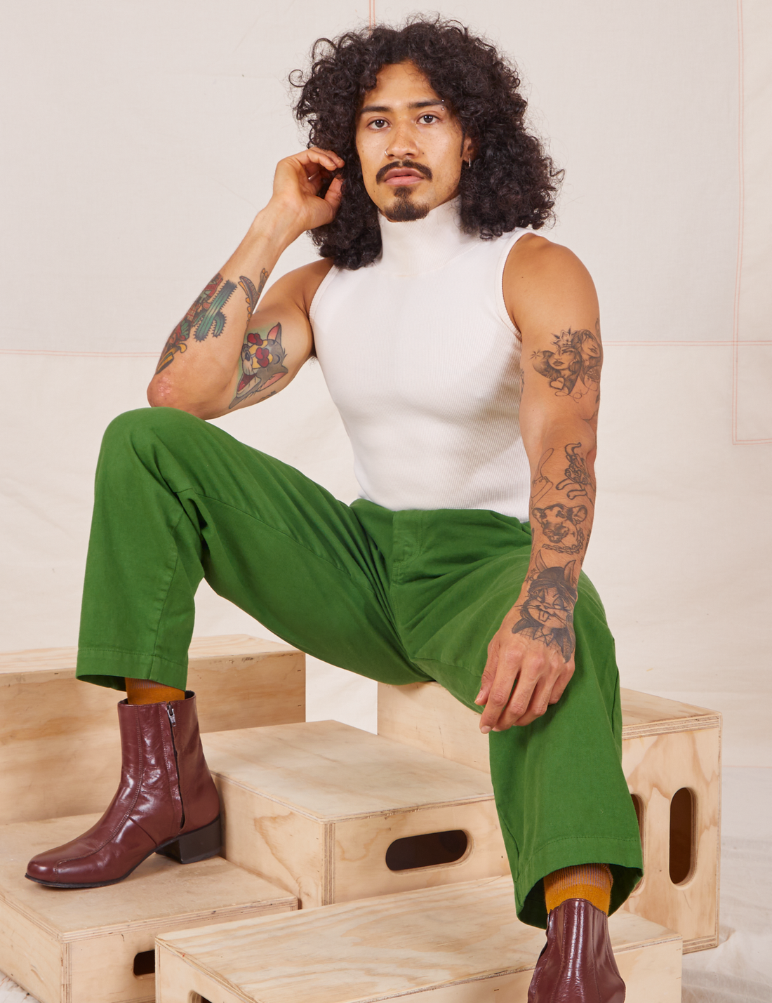 Jesse is sitting on a wooden crate wearing Heavyweight Trousers in Lawn Green and vintage off-white Sleeveless Turtleneck