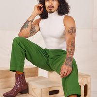 Jesse is sitting on a wooden crate wearing Heavyweight Trousers in Lawn Green and vintage off-white Sleeveless Turtleneck