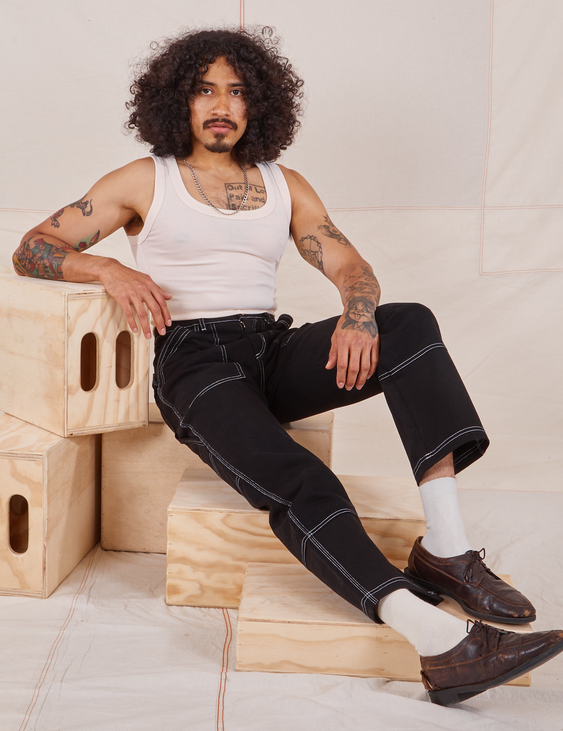 Jesse is sitting on a wooden crate. They are wearing Carpenter Jeans in Black and our vintage off-white Tank Top.