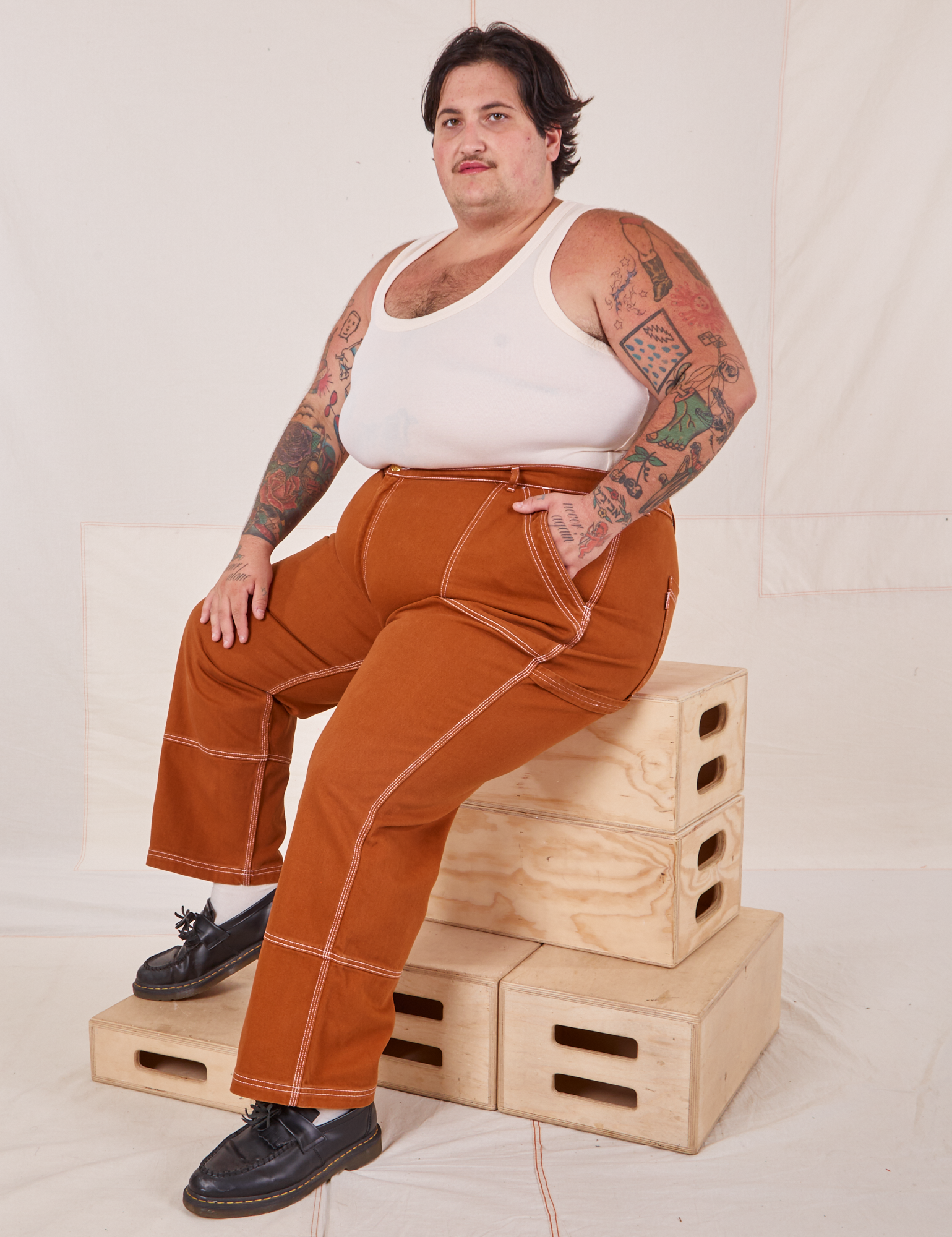 Sam is wearing Carpenter Jeans in Burnt Terracotta and Tank Top in vintage tee off-white. They are sitting on a stack of wooden crates.
