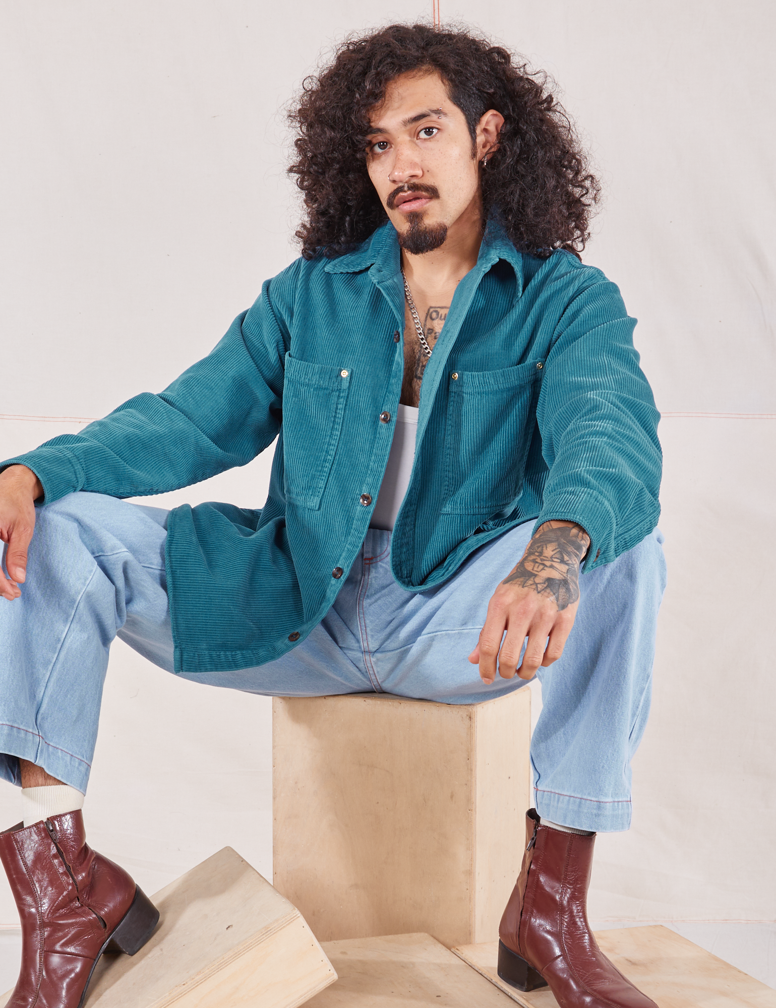 Jesse is wearing Corduroy Overshirt in Marine Blue and light wash Denim Trouser Jeans sitting on a wooden crate