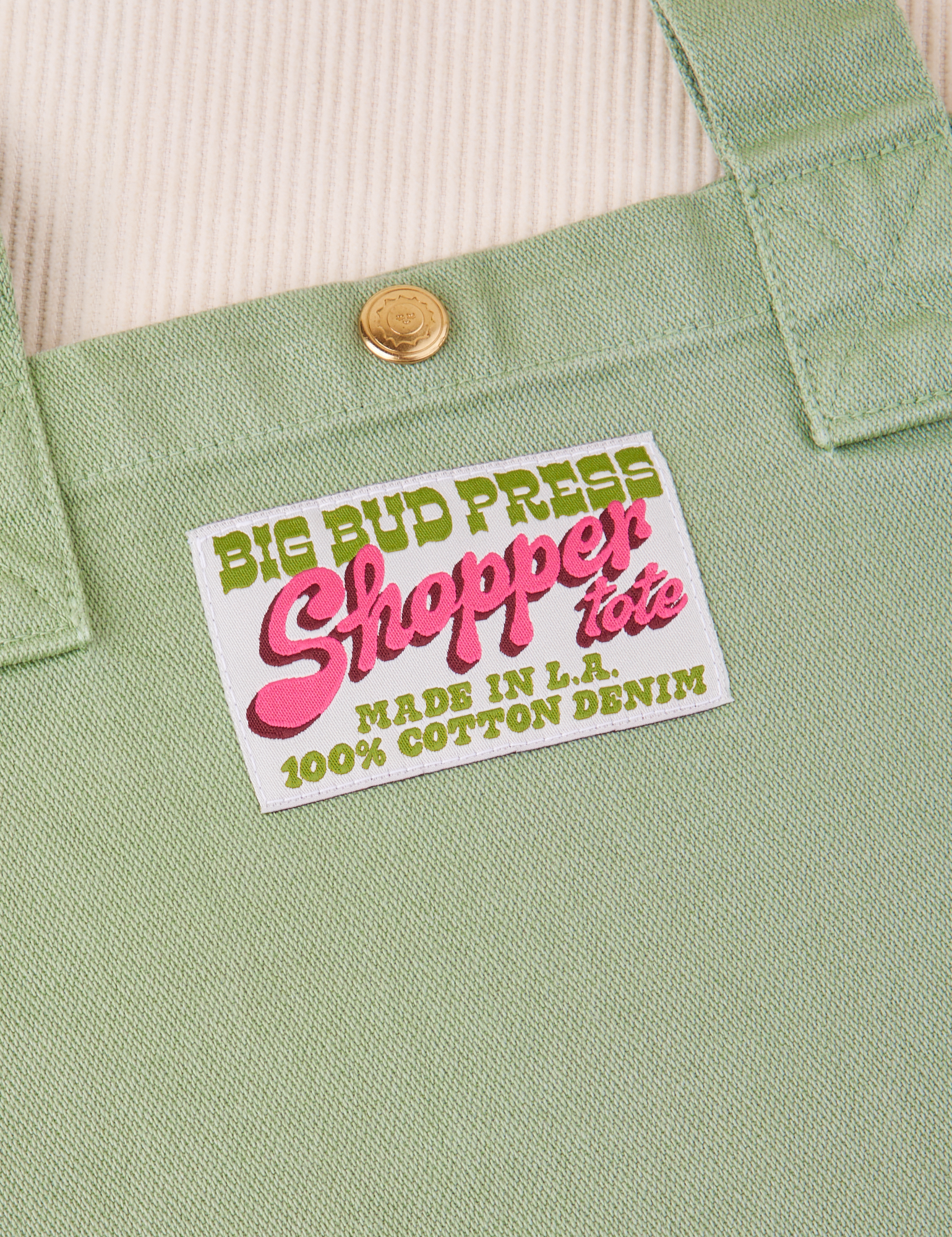 Sun Baby brass snap on Shopper Tote Bag in Sage Green. Bag label with green and pink text that reads &quot;Big Bud Press Shopper Tote, Made in L.A., 100% Cotton Denim&quot; on white background