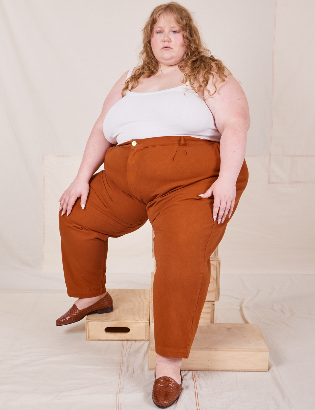 Catie is sitting on a stack of wooden crates wearing Heavyweight Trousers in Burnt Terracotta and vintage off-white Cropped Cami