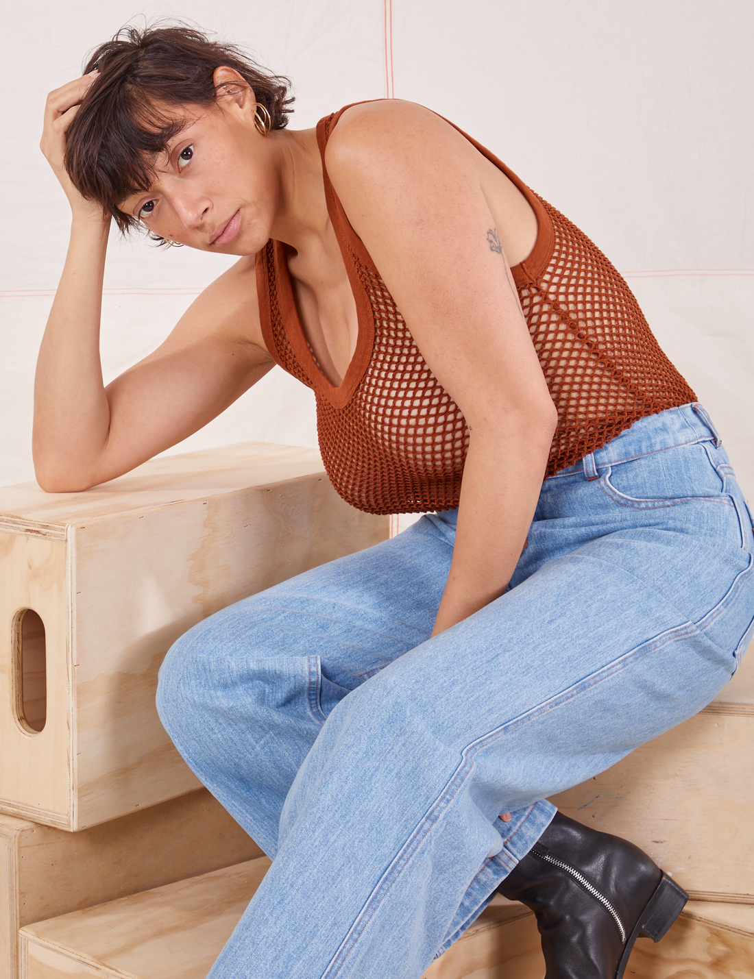 Tiara is sitting on a wooden crate wearing Mesh Tank Top in Burnt Terracotta and light wash Sailor Jeans
