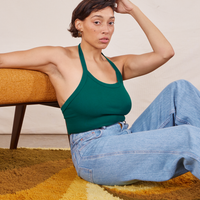 Tiara is sitting on a shaggy yellow rug, leaning on a vintage upholstered chair. She is wearing Halter Top in Hunter Green and light wash Sailor Jeans