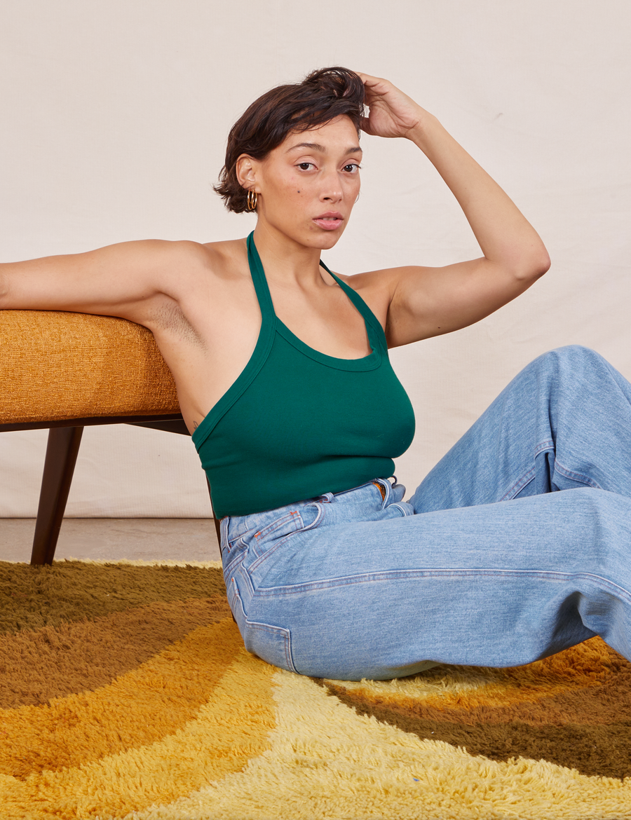 Tiara is sitting on a shaggy yellow rug, leaning on a vintage upholstered chair. She is wearing Halter Top in Hunter Green and light wash Sailor Jeans