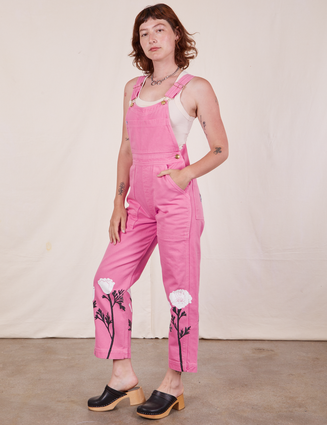 Angled view of California Poppy Overalls in Bubblegum Pink and vintage off-white Tank Top worn by Alex. She has her left hand on her left leg and the right hand in the pant pocket.