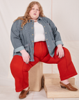 Catie is sitting on a wooden crate wearing Railroad Stripe Denim Work Jacket and paprika Western Pants