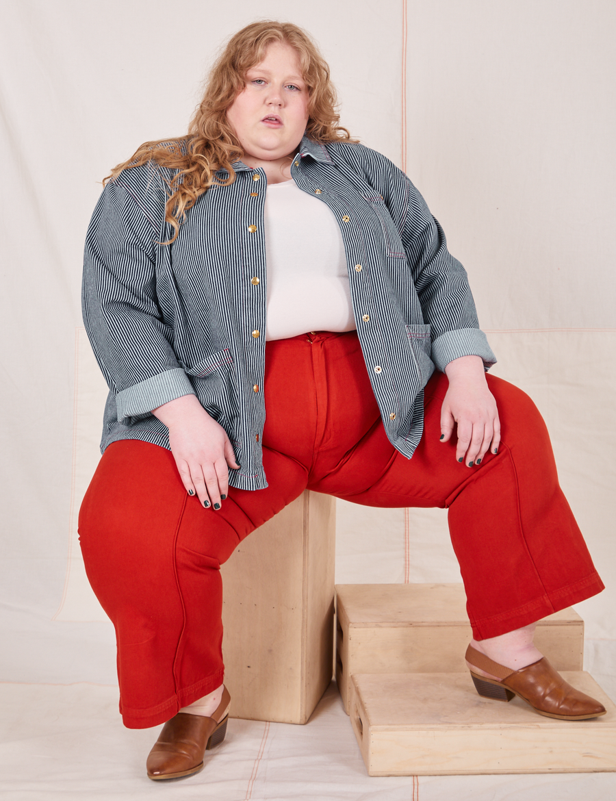 Catie is sitting on a wooden crate wearing Railroad Stripe Denim Work Jacket and paprika Western Pants
