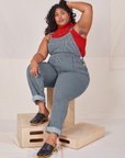 Morgan is sitting on a wooden crate wearing Railroad Stripe Denim Original Overalls and paprika Sleeveless Turtleneck