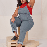 Morgan is sitting on a wooden crate wearing Railroad Stripe Denim Original Overalls and paprika Sleeveless Turtleneck