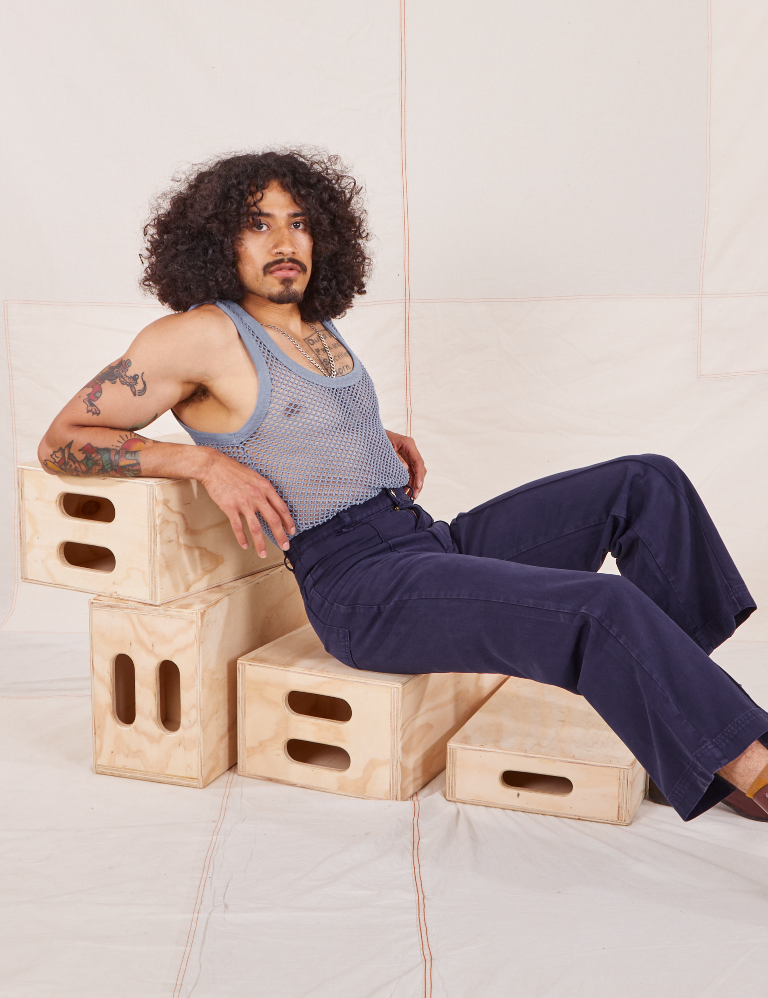 Jesse is sitting on a wooden crate wearing Mesh Tank Top in Periwinkle and navy Western Pants