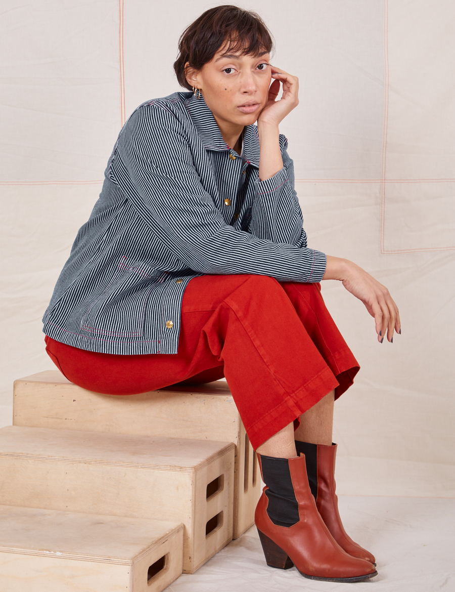 Tiara is sitting on a wooden crate wearing Railroad Stripe Denim Work Jacket and paprika Western Pants