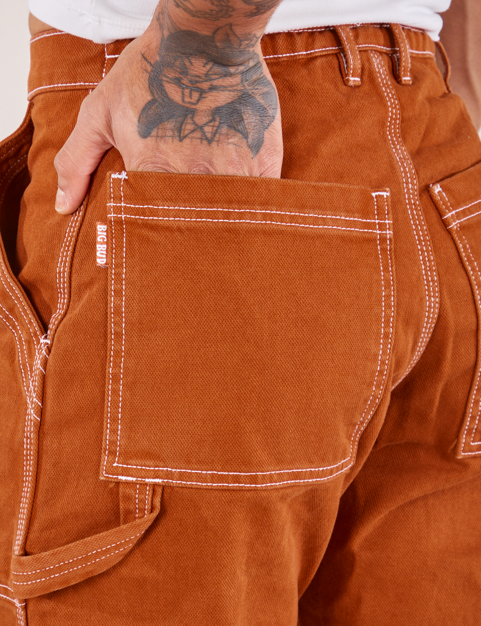 Carpenter Jeans in Burnt Terracotta back pocket close up. Jesse has their hand in the pocket.