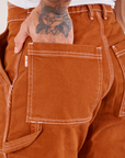 Carpenter Jeans in Burnt Terracotta back pocket close up. Jesse has their hand in the pocket.