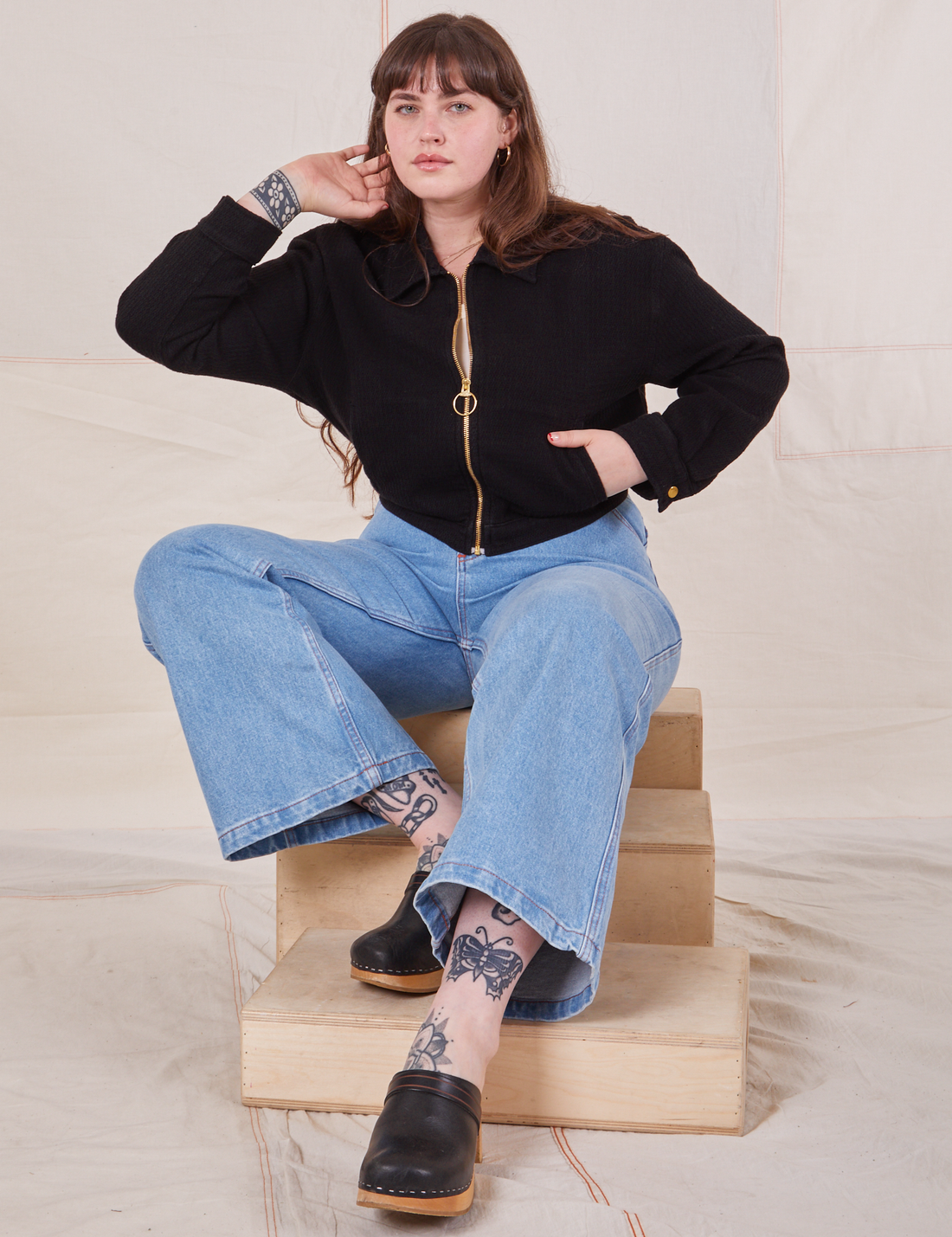 Sydney is sitting on a wooden crate wearing the Ricky Jacket in Basic Black and light wash Sailor Jeans