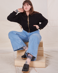 Sydney is sitting on a wooden crate wearing the Ricky Jacket in Basic Black and light wash Sailor Jeans