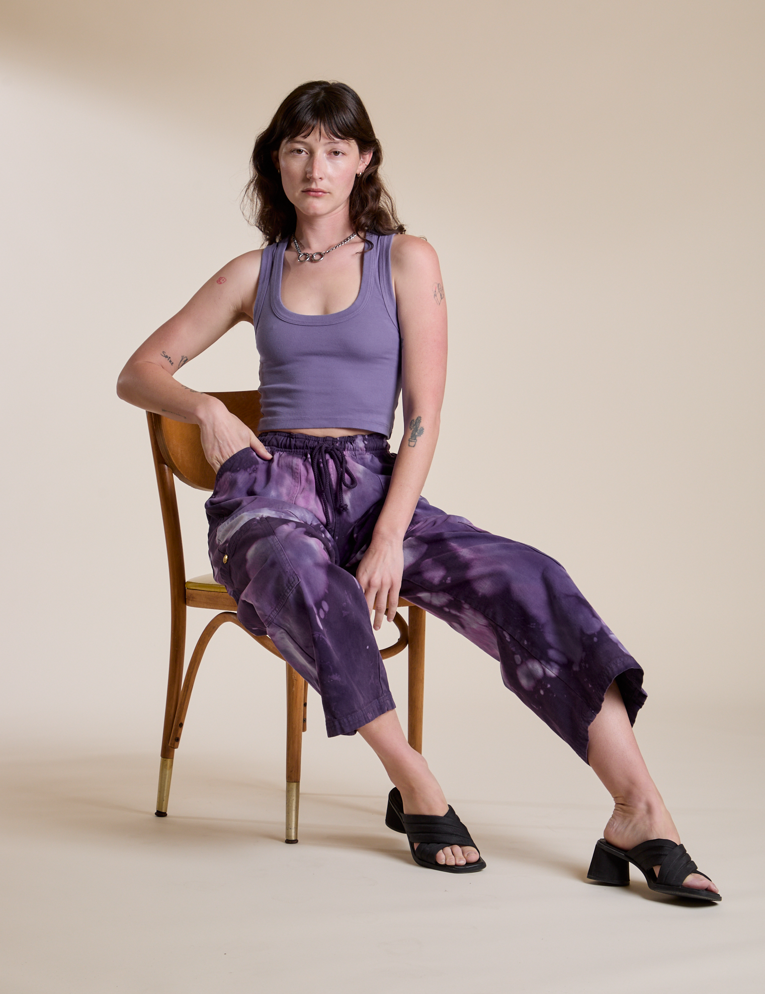 Alex is sitting in a chair and wearing Magic Waters Drop Pocket Pants in Dark Lavender paired with faded grape Cropped Tank