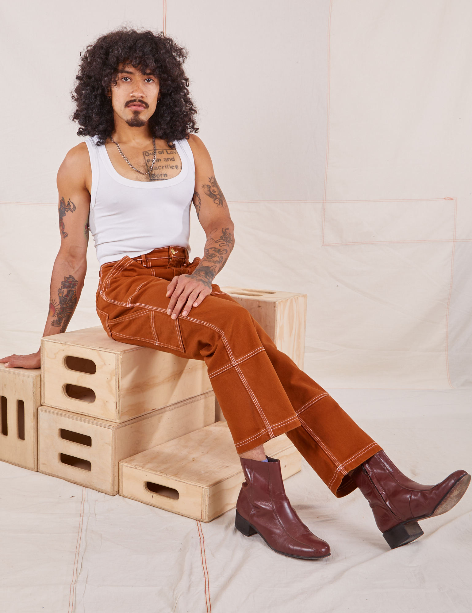 Jesse is sitting on a stack of wooden crates. They are wearing Carpenter Jeans in Burnt Terracotta and vintage off-white Cropped Tank Top