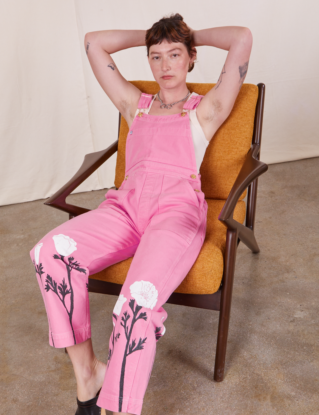 Alex is sitting on a vintage wooden chair with orange upholstery. She is wearing California Poppy Overalls in Bubblegum Pink and a vintage off-white Tank Top underneath.