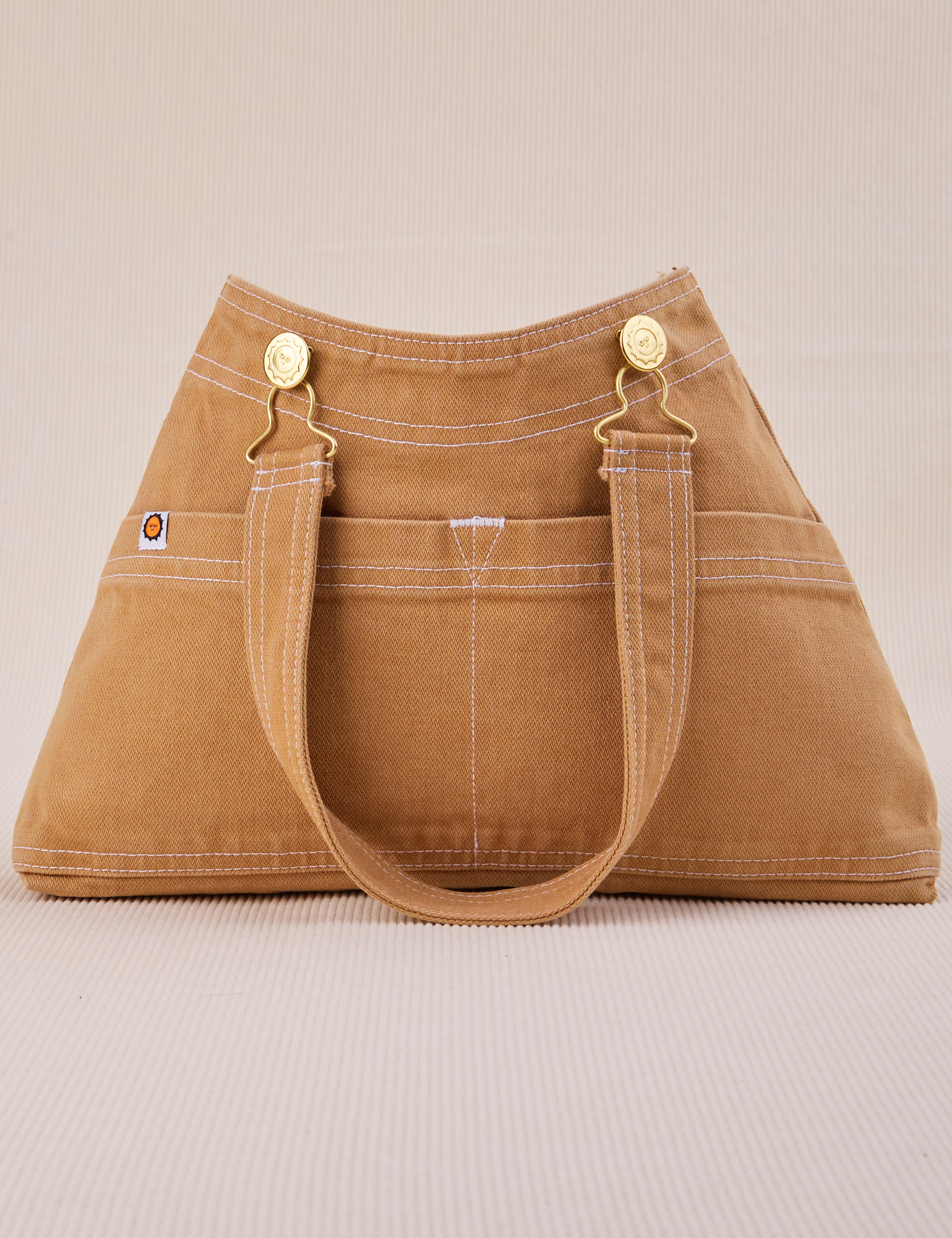 Overall Handbag in Tan with handle strap hanging down front of bag.