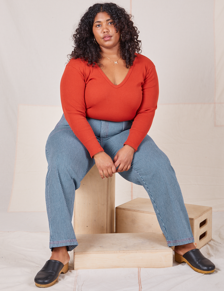 Morgan is sitting on a wooden crate wearing Railroad Stripe Denim Work Pants and a paprika Long Sleeve V-Neck Tee
