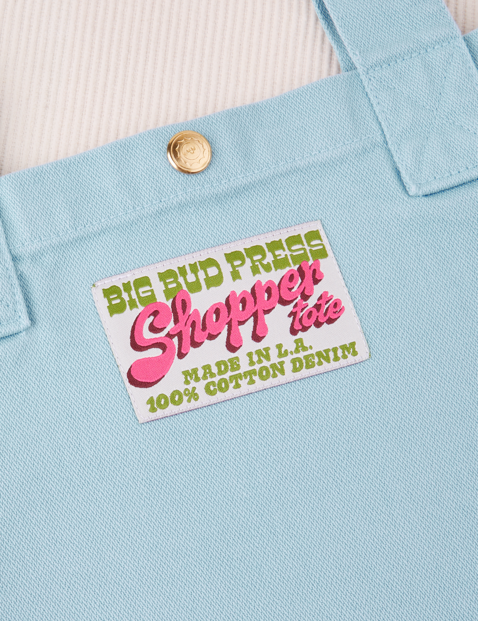 Sun Baby brass snap on Shopper Tote Bag in Baby Blue. Bag label with green and pink text that reads &quot;Big Bud Press Shopper Tote, Made in L.A., 100% Cotton Denim&quot; on white background
