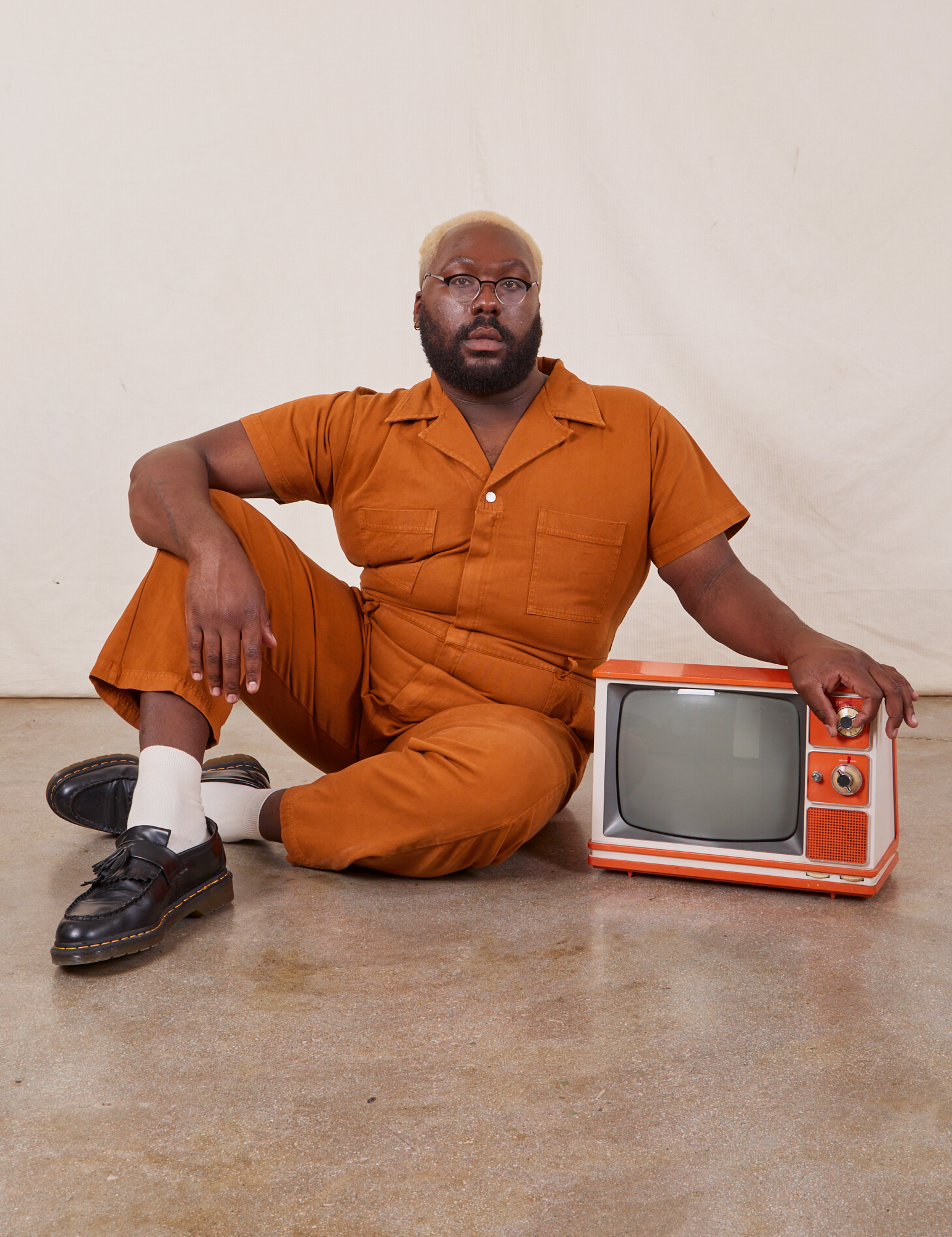 Elijah is wearing Short Sleeve Jumpsuit in Burnt Terracotta and sitting next to a small vintage tv.