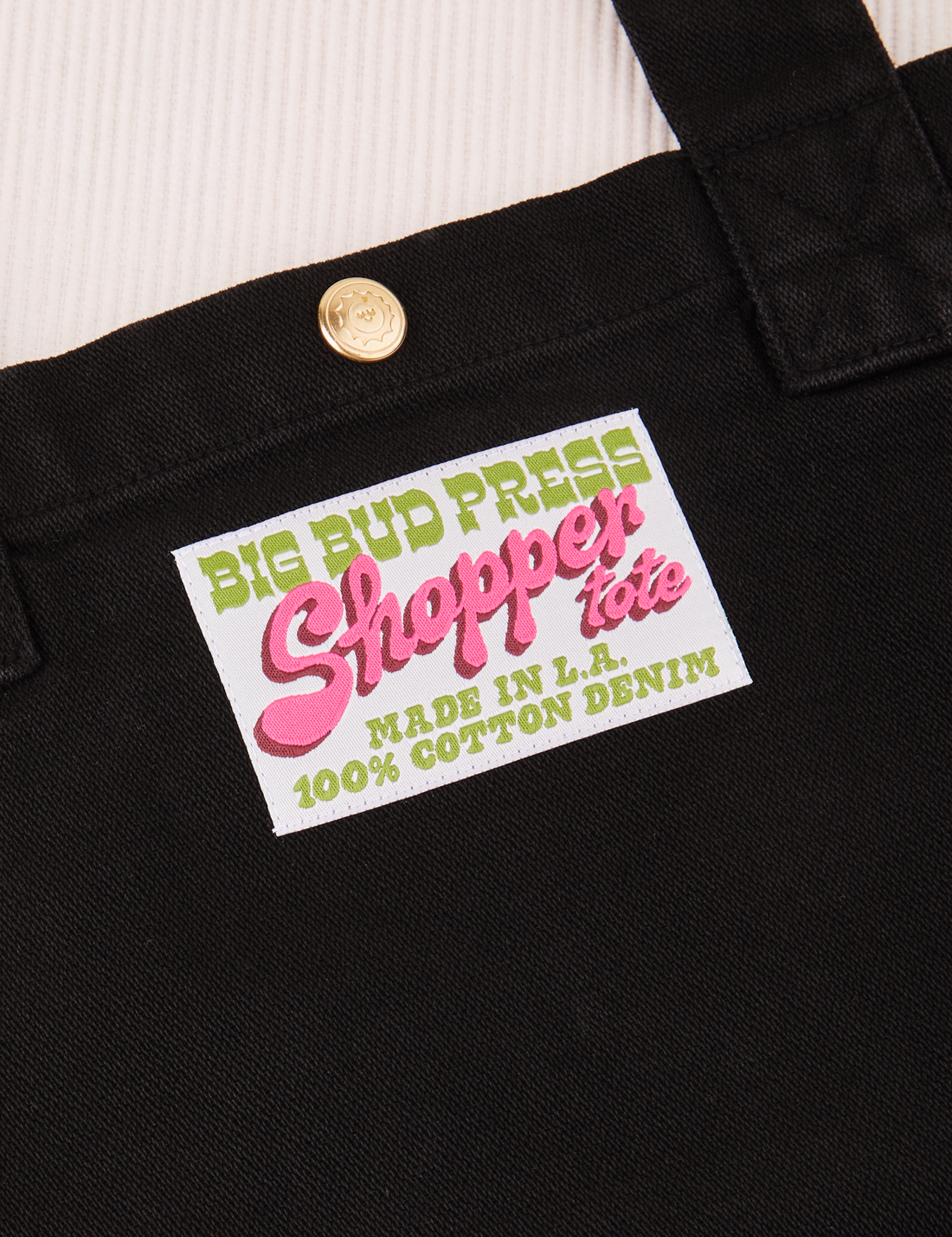 Sun Baby brass snap on Shopper Tote Bag in Black. Bag label with green and pink text that reads "Big Bud Press Shopper Tote, Made in L.A., 100% Cotton Denim" on white background