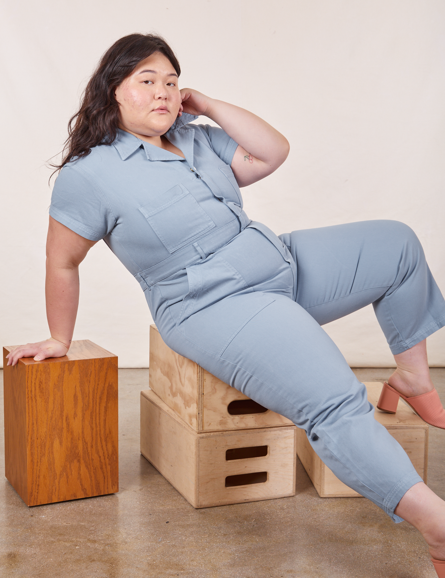 Ashley is wearing Petite Short Sleeve Jumpsuit in Periwinkle and sitting on a stack of wooden crates