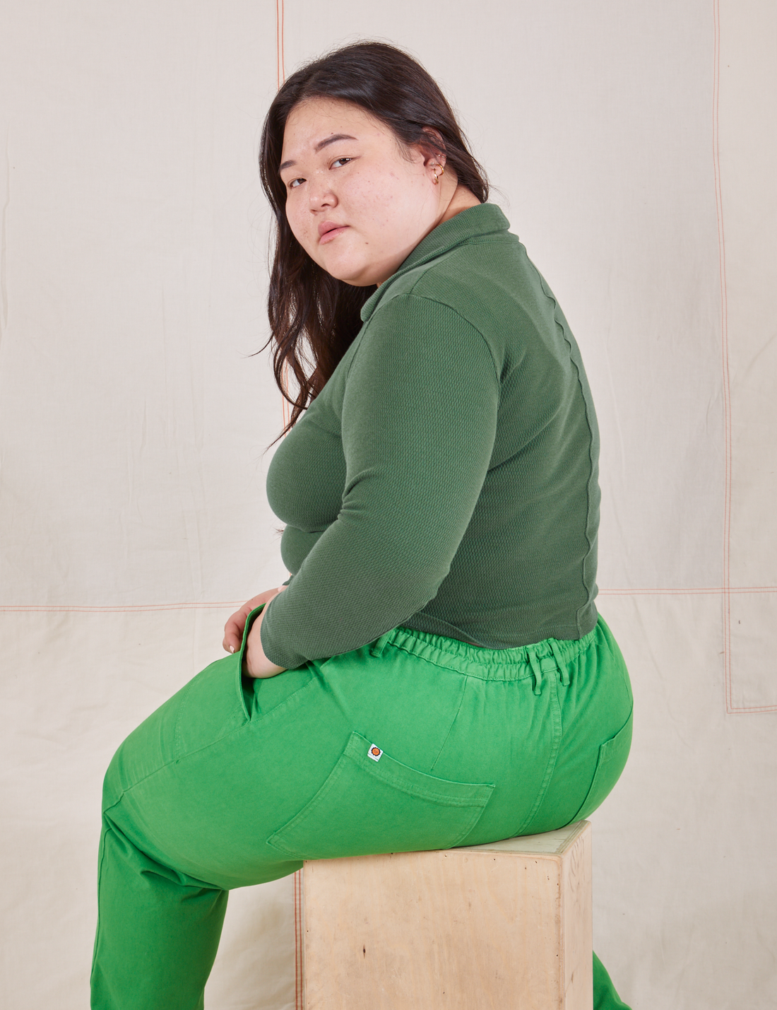 Long Sleeve Fisherman Polo in Dark Emerald Green side view on Ashley wearing kelly green Work Pants sitting on wooden crate